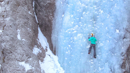 AERIAL: Unrecognizable woman climbs up a beautiful frozen waterfall in the Alps.