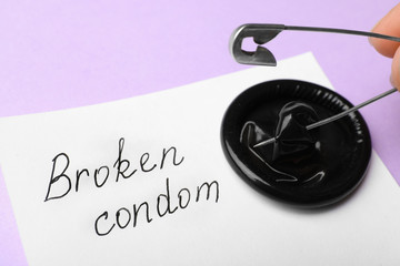 Woman piercing condom with pin on violet background, closeup. Safe sex