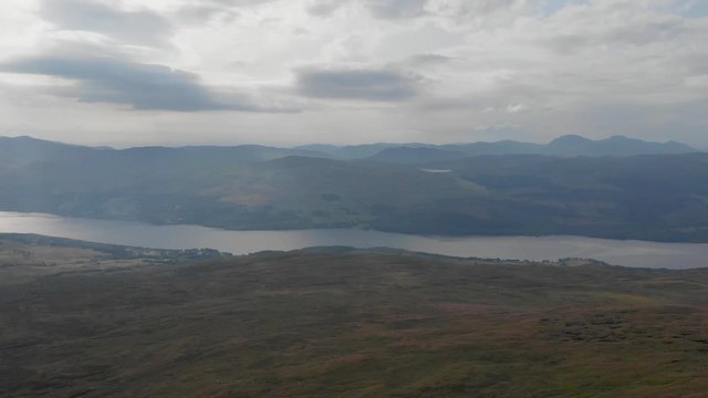 An aerial sideway left to right footage of a grassy mountain plateau with a huge lake and mountain range in the background under a cloudy sky	