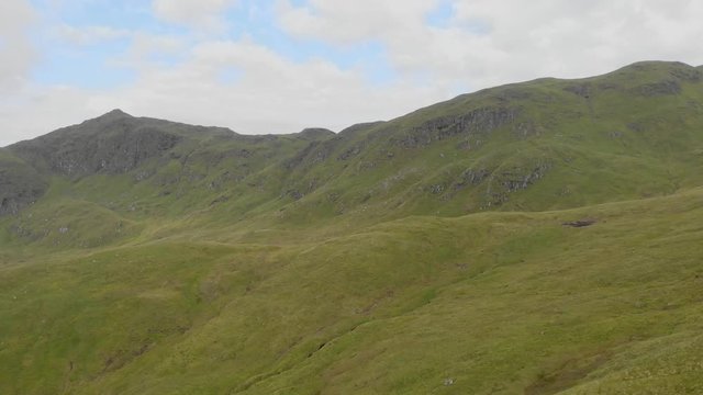 An aerial forward footage of a grassy mountain slope and rocky summit under a blue sky and white clouds