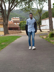 Fototapeta na wymiar Hombre joven negro de Senegal quitándose la chaqueta mientras camina con camisa clara arrugada , pantalones vaqueros estrechos y zapatillas blancas.