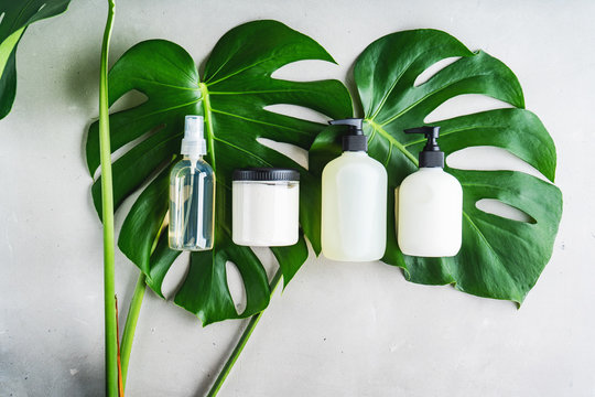 Cosmetic set of blank label bottles for mockup packaging of skincare product cream, serum, oil, shampoo, conditioner, perfume on grey background with green leaves. Natural beauty product concept.