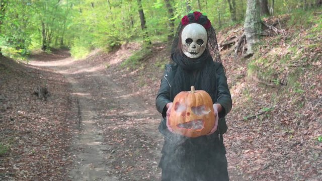 Woman close on with creepy skull mask in the autumn woods, holding a carved Halloween  smoky pumpkin.  On background path with  fall leaves. Concept about Halloween holidays and Day of the dead.