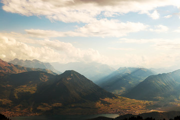 Valley illuminated by the sunset, sun rays visible. High mountains and clouds in the background. Below the lake with small villages; very warm and relaxing colors.