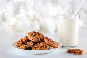 Closeup to delicious handmade chocolate chip cookies on a white plate and a glass of fresh milk on Christmas day. The lights are on decorating the white Christmas tree. Christmas holidays concept
