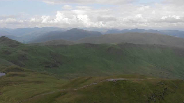 An aerial forward footage of a grassy mountain rocky summit with small pond and mountain range in the background under a cloudy sky