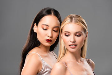 elegant blonde and brunette women in satin dresses looking down isolated on grey