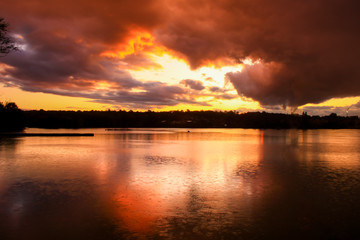 Dramatic sky at sunset over water with a pier in the foreground in the middle of a lake. Silhouette of forest in the background. 