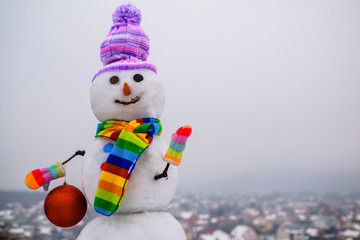 Snowman holds new year ball. Snow man in winter hat. Christmas background with snowman. Funny snowman in stylish hat and scarf. Smiling snow man with Christmas toy. Merry Christmas and Happy new year.