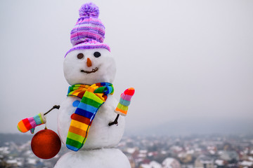 Obraz premium Snowman holds new year ball. Snow man in winter hat. Christmas background with snowman. Funny snowman in stylish hat and scarf. Smiling snow man with Christmas toy. Merry Christmas and Happy new year.