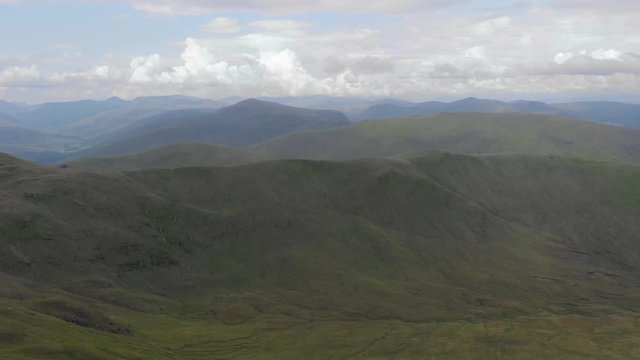 An aerial backward footage of a grassy mountain rocky summit with small pond and mountain range in the background under a cloudy sky