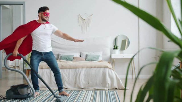 Joyful guy in superman costume red cape and mask cleaning carpet with vacuum cleaner with raised arm doing housework alone. People and super heros concept.