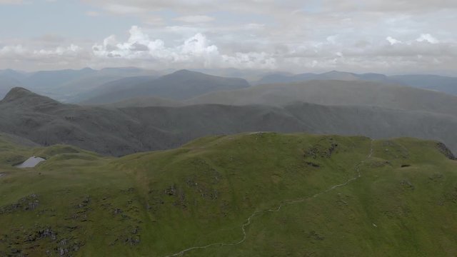An aerial forward footage of a grassy mountain rocky summit with small pond and mountain range in the background under a cloudy sky