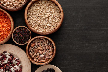 top view of brown bowls with raw beans, lentil, oatmeals and quinoa on dark wooden surface with copy space