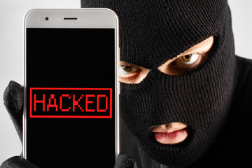 Male hacker thief in mask and gloves holding stolen smartphone.