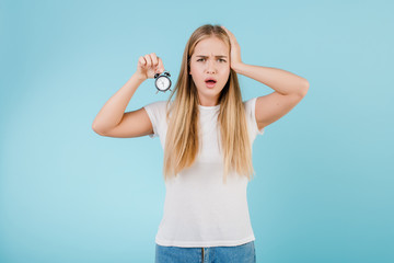 beautiful young blonde woman with alarm clock showing time isolated over blue