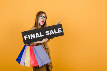 hipster woman in glasses and coat with final sale sign and colorful shopping bags isolated over yellow