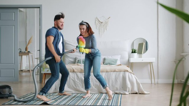 Woman and man are singing in duster and playing the guitar on vacuum cleaner in bedroom having fun during clean-up. People, housekeeping and happiness concept.