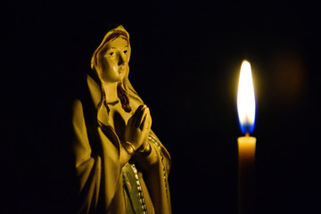 Statuette of the Virgin Mary and a lit church candle against dark background. Close-up. Selective...