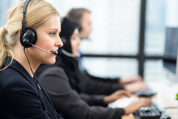 Beautiful Caucasian female call center with headset talking to customer in office. The girl working as technical service support. Seen from woman side with colleagues muslim girl and Caucasian man.