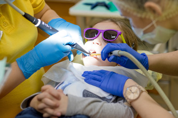 Dentist performing dental filling procedure to preteen girl in pediatric dental clinic. Doctor removing caries using high-speed dental drill. Calm child is sitting in a dental chair holding mouth open