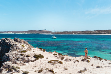 Woman in bikini, on a bright summers day at the beach looking out admiring the view of the stunning ocean and a white boat, at a bay on Rottnest Island, Western Australia.