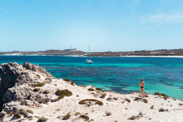 Fototapeta premium Woman in bikini, on a bright summers day at the beach looking out admiring the view of the stunning ocean and a white boat, at a bay on Rottnest Island, Western Australia.