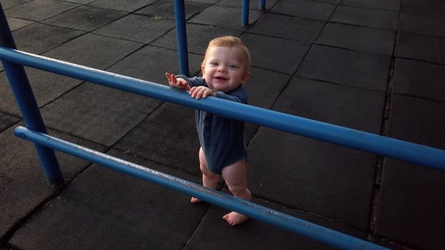 Happy toddler boy stands alone on rubber playground mat leaning against blue jungle gym looking over bars. Faces toward, looks up, and smiles at camera. calm peaceful scene during golden hour sunset.