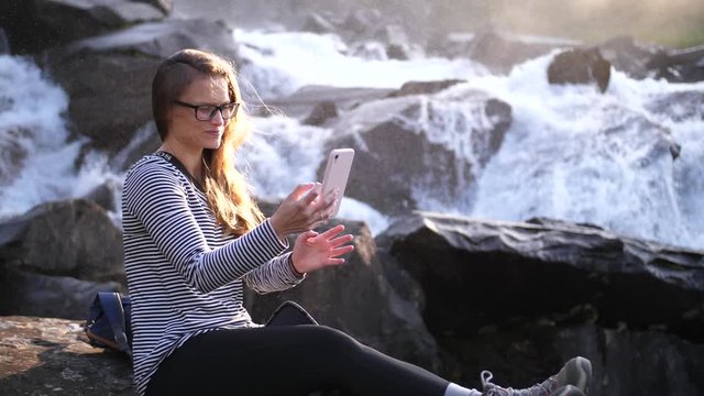 A young, brunette woman sitting by herself on a rock with wind blowing in her hair while taking a selfie photo of herself with her phone. Big river rushing down behind her with the sunset shining.