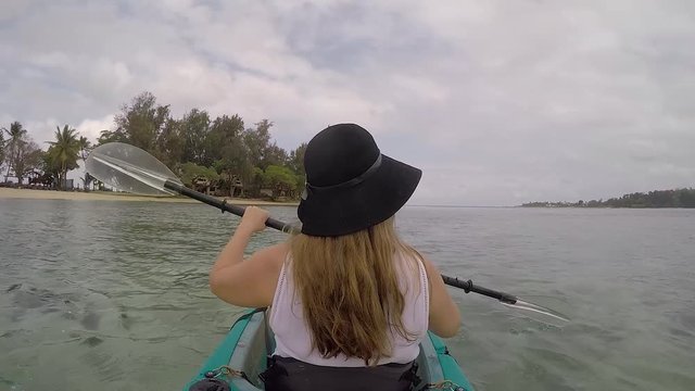 Girl with long blonde hair wearing a black hat paddling a kayak near a tropical island