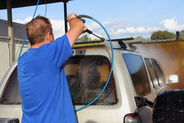A man washes the railings on roof of a silver SUV car with water jet from a high pressure sink washer on a summer day outdoor