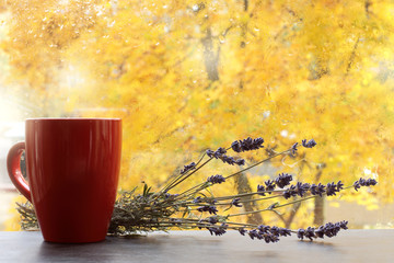 red mug and a bouquet of lavender on a table opposite a window with raindrops and yellow trees....