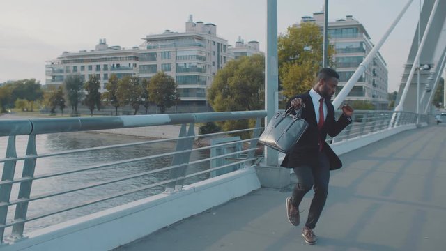 Happy african american businessman in casual suit with briefcase, walking walking by street of downtown funny dancing to the rythm. Young black business guy