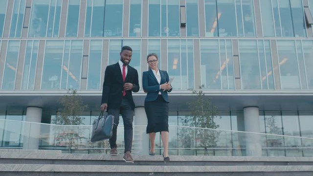 Two employees, delegates or lawyers walk on stairs from business center. Colleagues finished work day in their modern glass office. Man and woman in formal suits