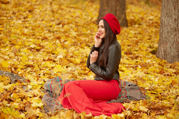 Outdoor shot of young attractive cute girl in red beret, trousers and leather jacket, sitting near trees on yellow leavesin autumn park with smart phone in her hand, talking with her boyfriend.