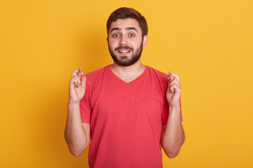 Lifestyle and people concept. Image og attractive guy waiting for special moment, young bearded man wearing red casual t shirt, keeping fingers while standing against yellow studio background.