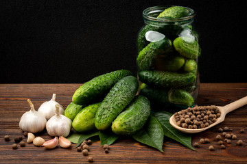 Fresh cucumbers in glass jar, garlic and black pepper on dark wooden background.
