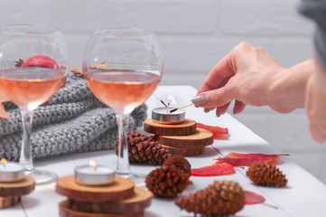 Small burning candles, two glasses with rose wine, cones, dry red leaves, gray scarf knitted on a white wooden table. Girl lights candles on the table. Romantic evening. Hello, Autumn. Cozy autumn bac