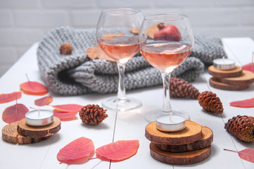 Small candles, two glasses with rose wine, cones, dry red leaves, a gray scarf knitted on a white wooden table. Hello, Autumn. Cozy autumn background.