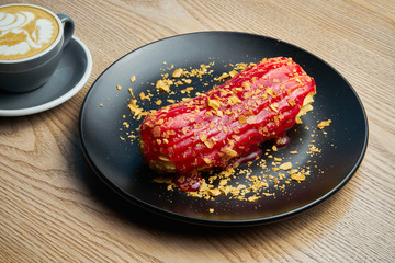 Appetizing and large eclair with glaze of raspberry on top and fir cream inside on a black plate. Sweets and dessert for coffee and tea.