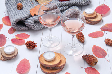 Small candles, two glasses with rose wine, cones, dry red leaves, a gray scarf knitted on a white wooden table. Hello, Autumn. Cozy autumn background.
