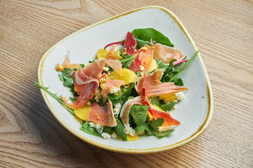 Close up view on tasty Light salad with prosciutto, a mix of greens, pineapple and blue cheese in stylish grey bowl.