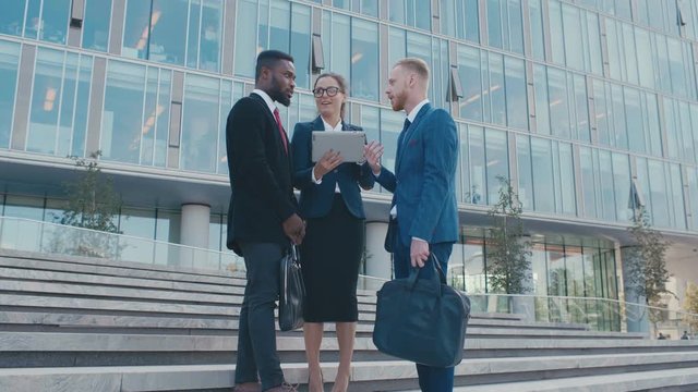 Group of business people wearing formal suits one woman and two men standing outside and talking. Partners and colleagues using looking tablet PC