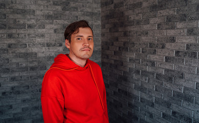 Obraz na płótnie Canvas A young handsome man in a red hoodie with bristles poses on a gray brick background.