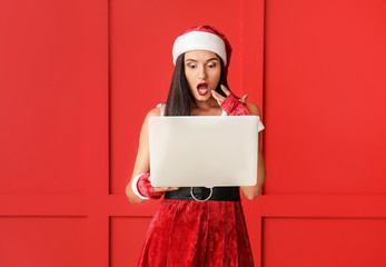 Surprised young woman in Santa Claus costume and with laptop on color background