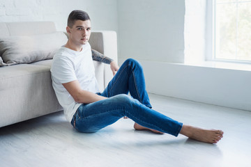 Young man in white t-shirt and jeans sits on the floor near the sofa in modern interior room