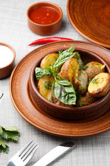 Fried potato in brown clay bowl close-up. Front view of plate with fried potato and basil with sauces and fork with knife. Focus on fried potato. Grey background. vertical photo