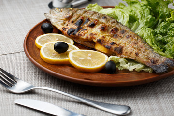 Grilled trout in brown clay bowl close-up. Front view of plate with grilled trout garnishe with salade, lemon and olives. Focus on fried fish. Grey background. horizontal photo