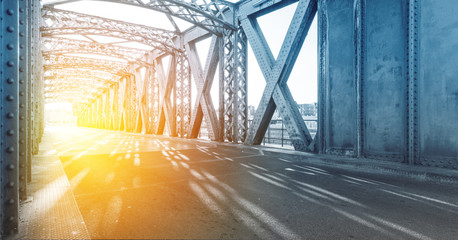 Asphalt road under the steel construction of a bridge in the city on a sunny day. Evening urban scene with the sunbeam in the tunnel. City life, transport and traffic concept.	