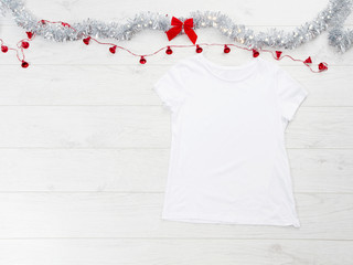 Close up white blank template t shirt with copy space and Christmas Holiday concept. Top view mockup t-shirt and red bow on white wooden background. Happy New Year decorations accessories. Xmas outfit